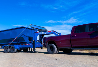 Blue roll-off dumpster container on a black Gridko gooseneck trailer, hitched to a maroon dually pickup truck on a dirt lot.