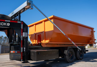 Orange roll-off dumpster container being loaded onto a black Gridko bumper pull trailer via its hydraulic tilt arm, parked in a lot with a chain-link fence and trees in the background.